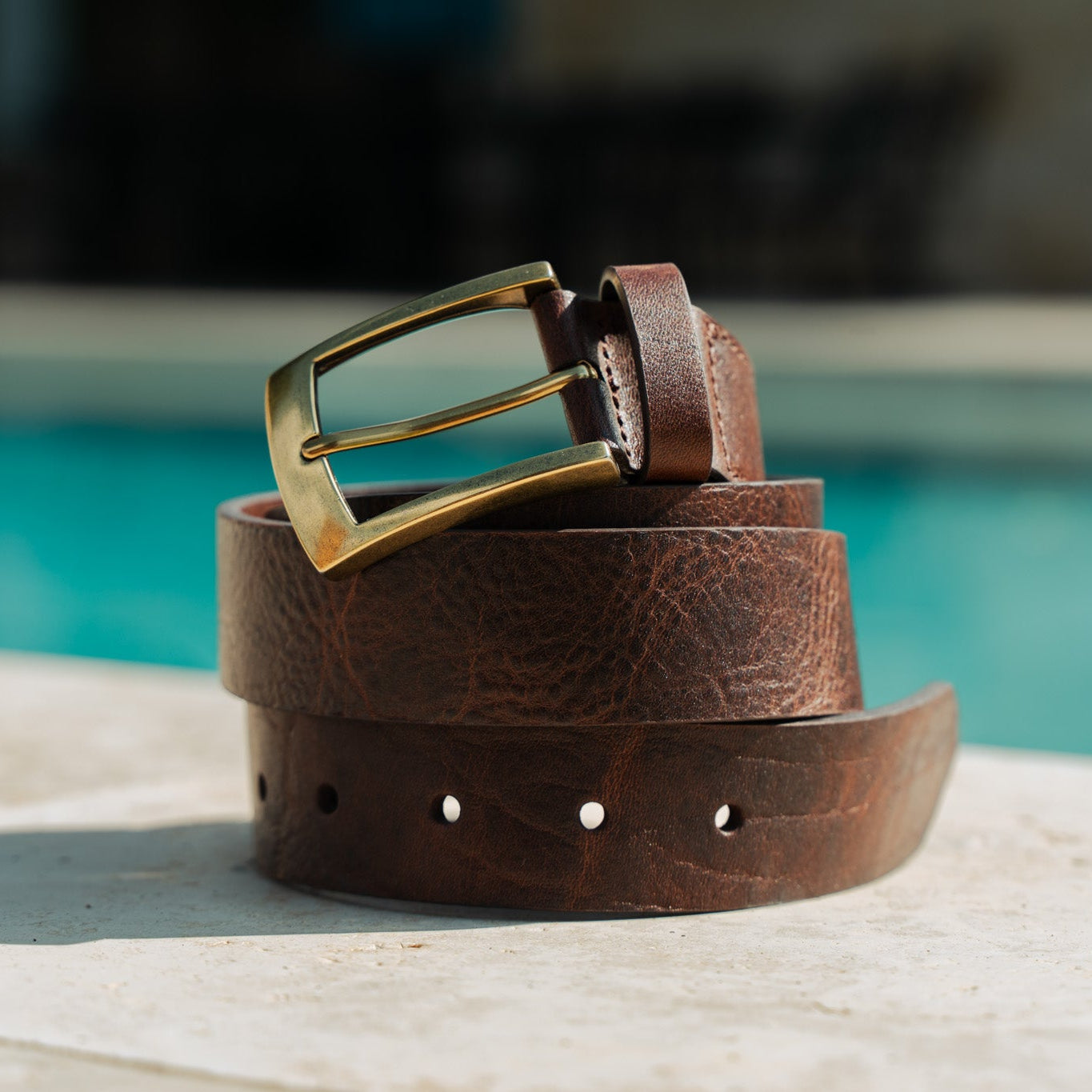 El Cuero full-grain leather belt coiled by a pool — aged brown leather with antique brass buckle showing natural patina
