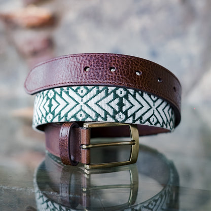 Tikal handwoven Maya belt coiled on a reflective glass surface — forest green and cream geometric pattern with full-grain leather ends and antique brass buckle