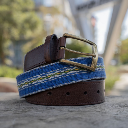Chapín handwoven Maya belt coiled on a stone surface — royal blue with white, green, and yellow stripe pattern, full-grain leather ends and antique brass buckle