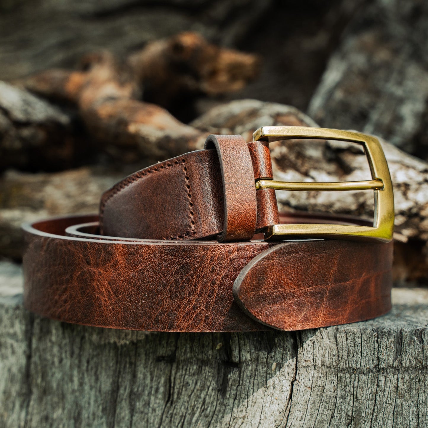 El Cuero full-grain leather belt resting on driftwood and rocks — rich brown patina with antique brass buckle and hand-stitched keeper
