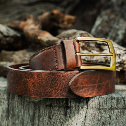 El Cuero full-grain leather belt resting on driftwood and rocks — rich brown patina with antique brass buckle and hand-stitched keeper