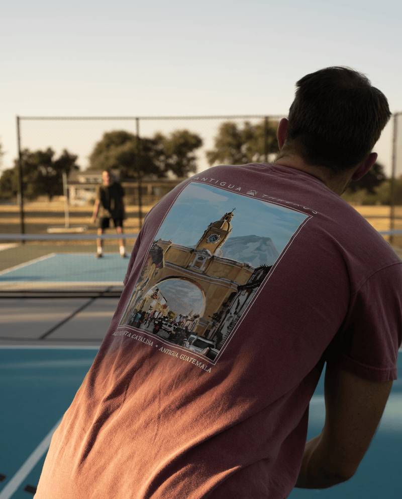 Person wearing a red Antigua Threads t-shirt featuring a large illustration of the Arco de Santa Catalina, playing pickleball outdoors on a court with another player in the background