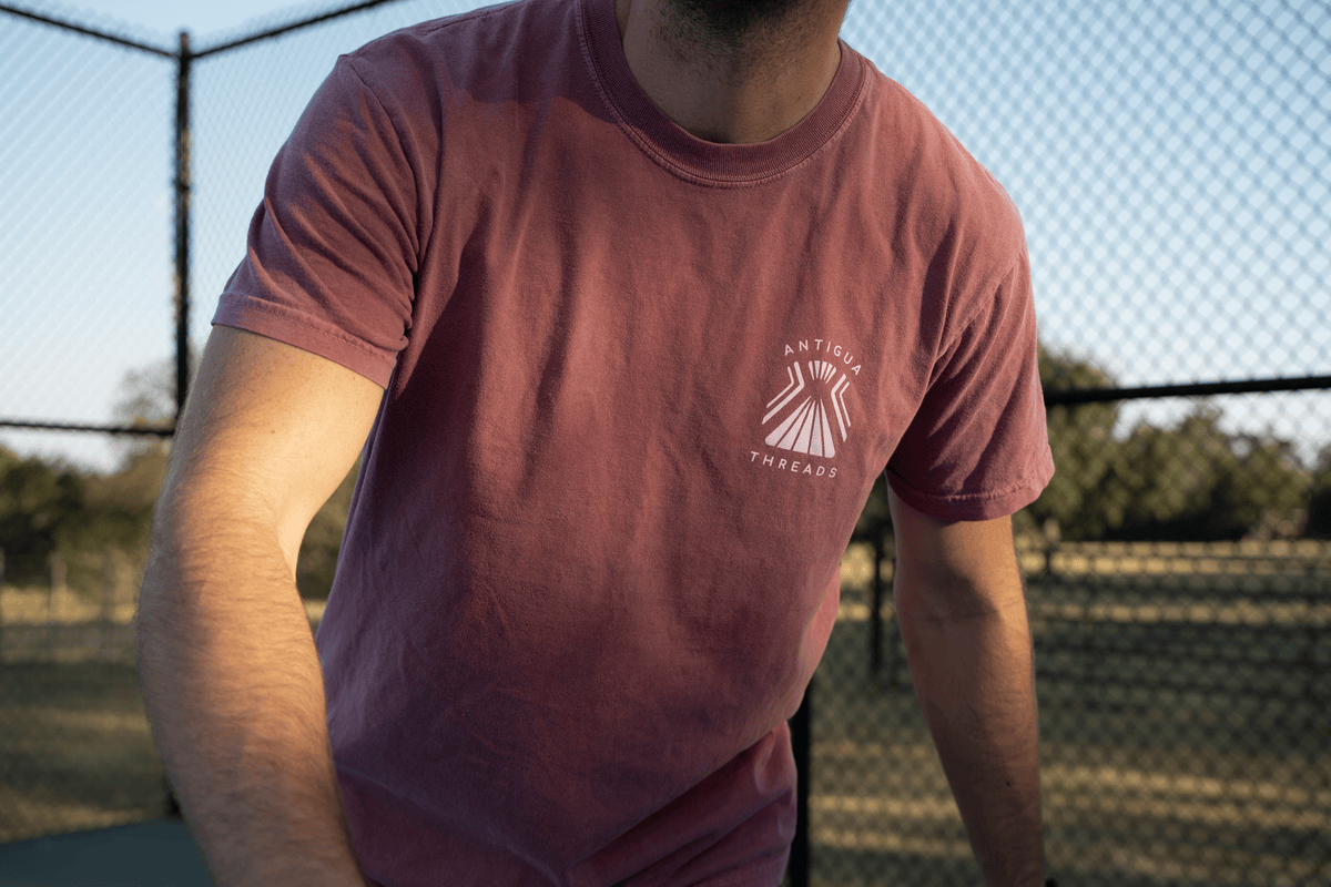 Person wearing a red Antigua Threads t-shirt with a white logo on the chest, playing pickleball outdoors on a court with a chain-link fence in the background